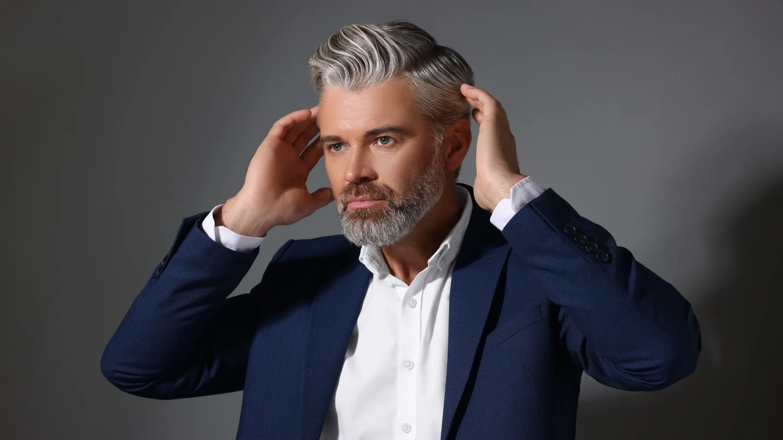 Man met grijs gemêleerd haar in een donkerblauw colbert strijkt met beide handen door zijn haar, symboolbeeld bij haartransplantatie